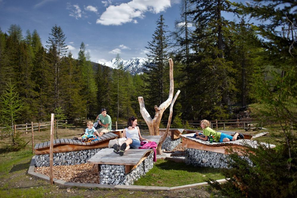 Eine Familie entspannt sich auf natürlichen Holzbänken, umgeben von Bäumen und Bergen unter einem blauen Himmel.