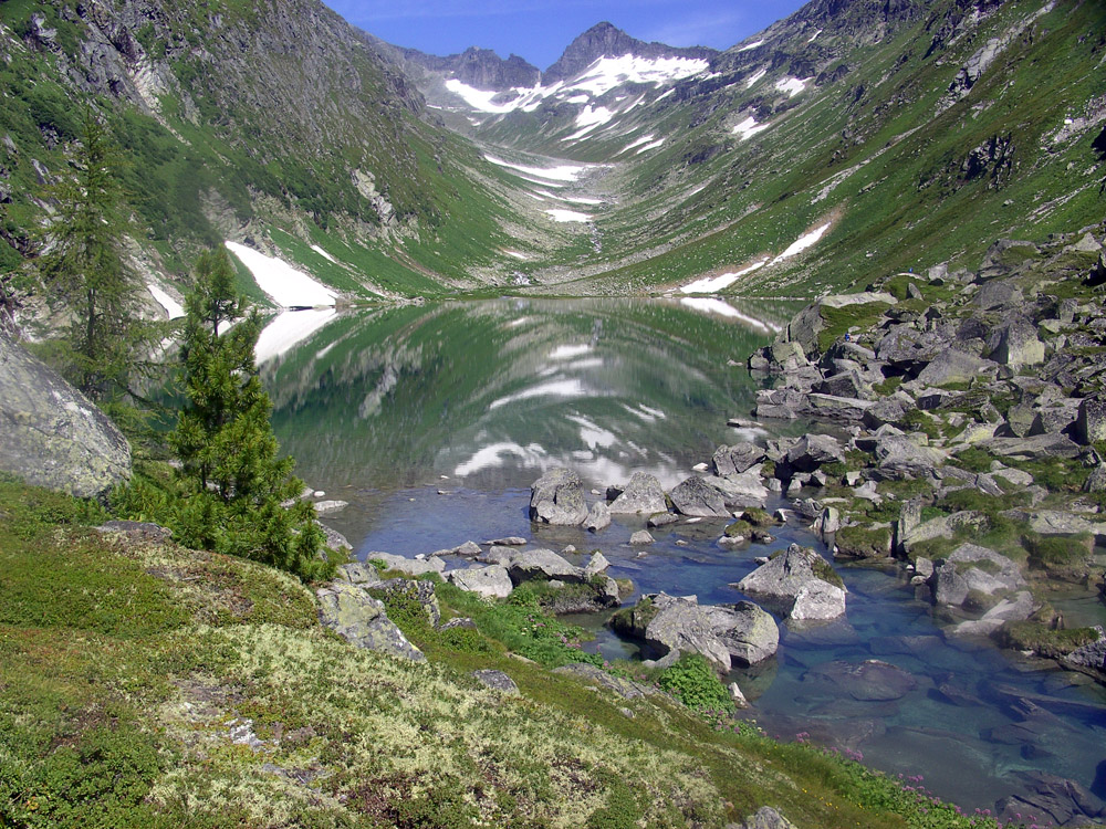 In einem ruhigen Bergsee spiegeln sich die umliegenden grünen Hügel und schneebedeckten Gipfel, umrahmt von felsigem Terrain und üppigem Grün.
