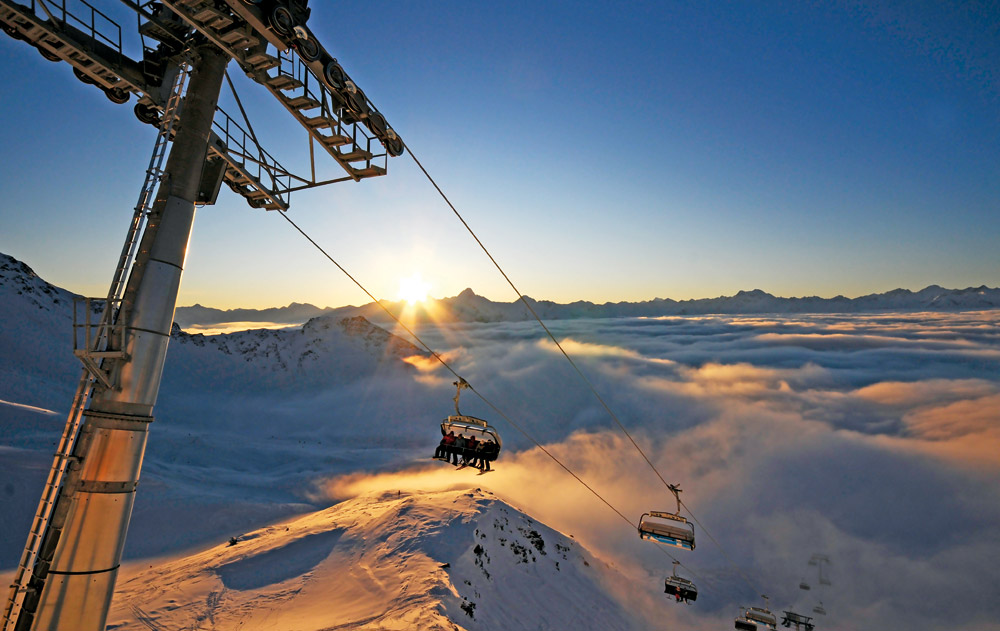Ein Skilift fährt schneebedeckte Berge hinauf, während die Sonne aufgeht, die Wolken darunter beleuchtet und die Landschaft in ein goldenes Licht taucht.