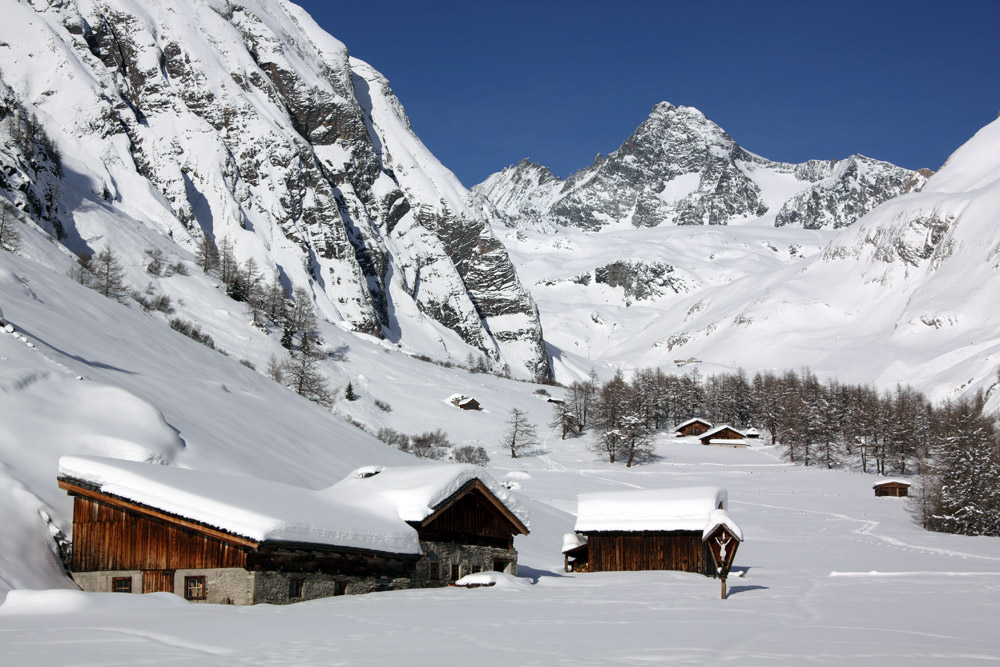 Schneebedeckte Almhütten in einem ruhigen Tal, umgeben von majestätischen Bergen unter einem klaren blauen Himmel.